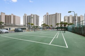 Una cancha de tenis con red, rodeada por una valla, junto a un estacionamiento con coches.