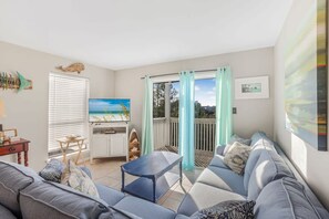 Un salon avec un canapé, une petite table et un téléviseur. Il y a un balcon d’où l’on peut admirer le ciel et les arbres.