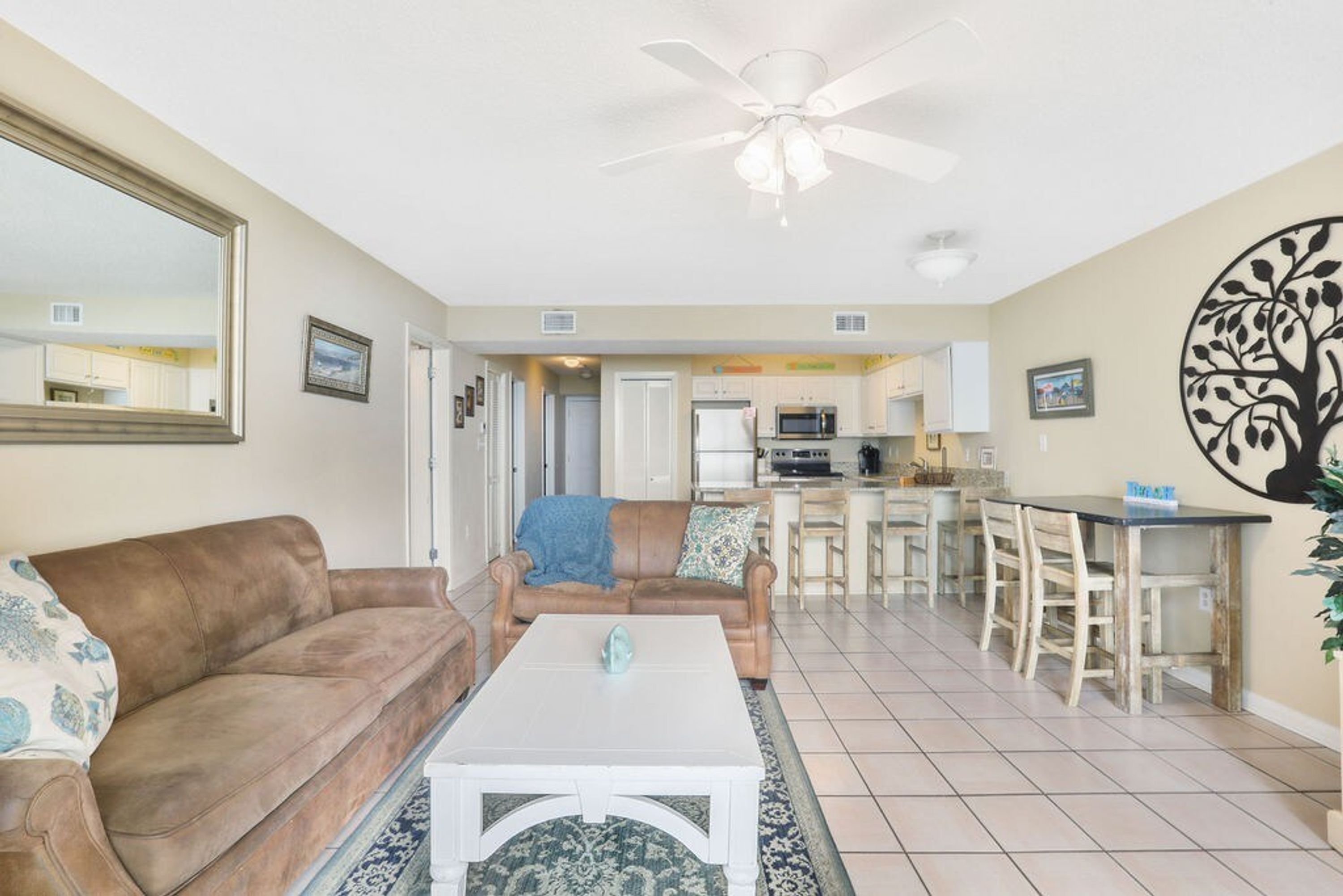 A living room with a brown leather couch, a white coffee table, and a ceiling fan. There is a kitchen in the background with a microwave and a dining area with chairs.