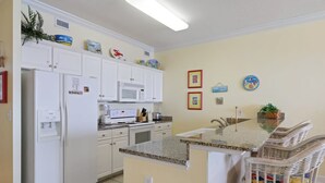 A kitchen with white cabinets, a microwave, a refrigerator, and a granite countertop. There are wicker chairs and a small island with a sink. - Regency Isle 711 2 BR 2 5 BA Sleep 8 Gulf-front (Orange Beach)