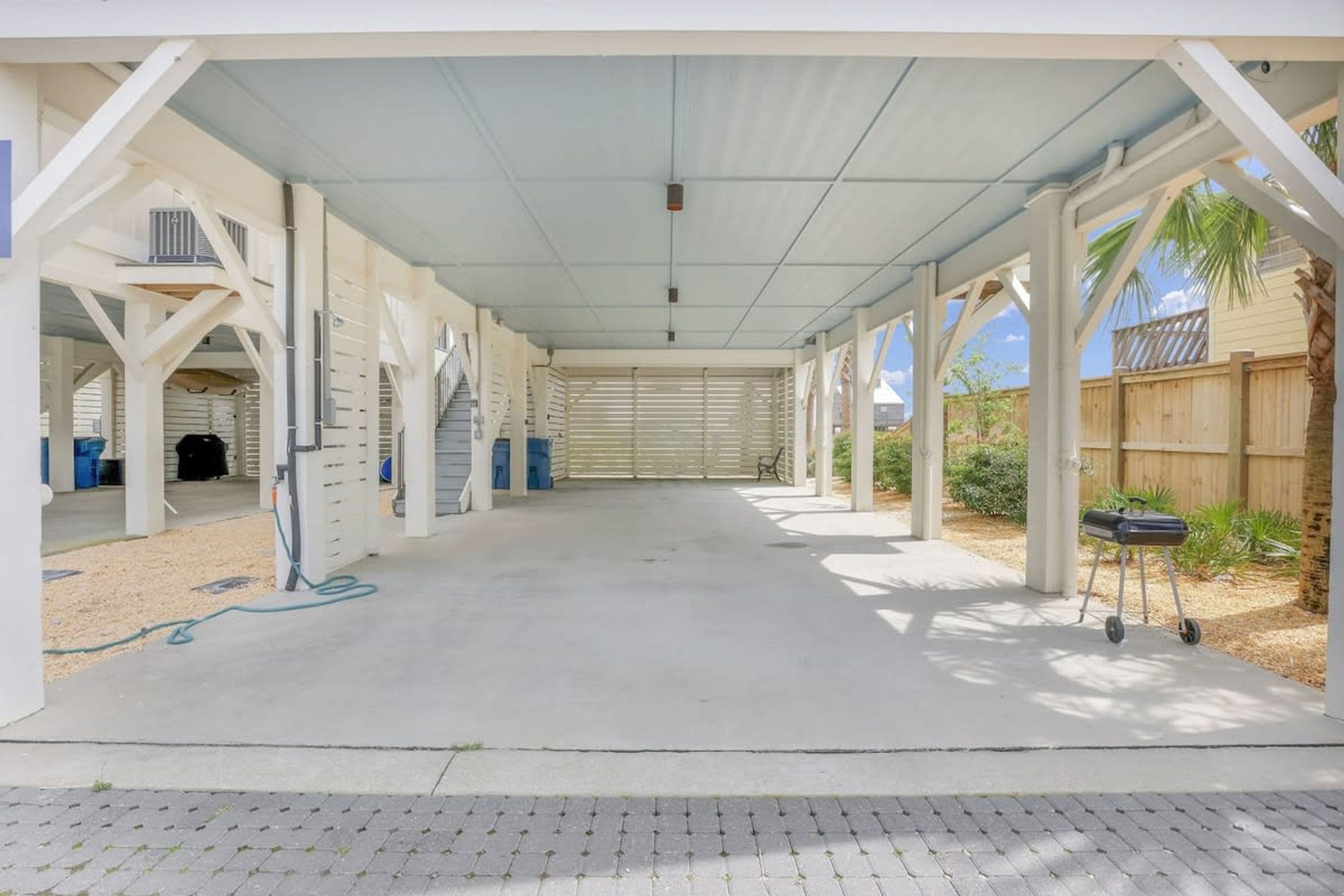 Un patio cubierto abierto con piso de concreto, vigas estructurales blancas y una cerca de madera al fondo.