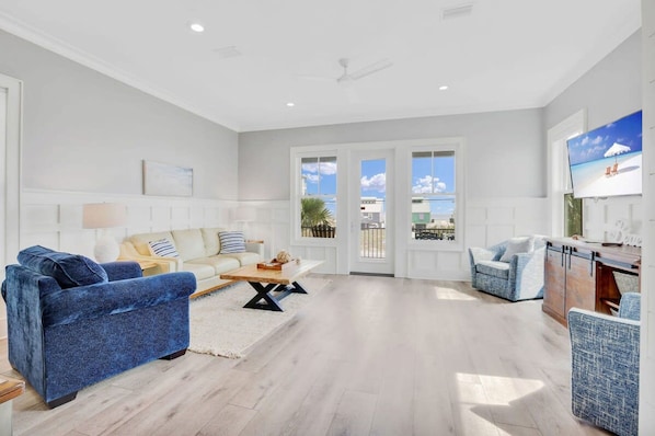 Un salon spacieux avec un canapé bleu, un canapé beige et un fauteuil bleu. On y trouve une table basse en bois et un téléviseur fixé au mur.