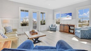 Un salón amplio con un sofá, un sillón y una mesa de centro. Hay ventanas grandes y un televisor montado en la pared.