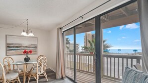 A balcony with a view of the ocean and buildings, a dining area with wicker chairs and a table, and a ceiling light fixture. - Lei Lani 218 Best of Beach Bay (Orange Beach)