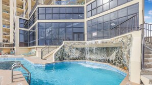 A swimming pool with a stone waterfall feature, surrounded by a multi-story building with balconies and large windows. - Phoenix II 2101-amenities Galore Beachfront (Orange Beach)