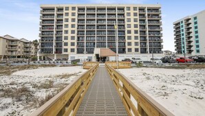 Un hotel de varios pisos con una entrada central, una playa de arena en primer plano y una pasarela de madera que conduce a la entrada.