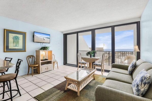 A spacious living area with a balcony, a dining table, and comfortable seating. - Island Wind West 177 Next to Hangout Pool Beach (Gulf Shores)