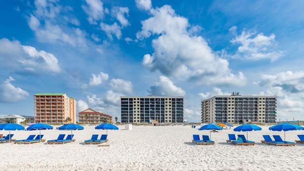 Una playa con hileras de tumbonas y sombrillas azules, un hotel con varios pisos y balcones, y un edificio más pequeño con techo rojo.