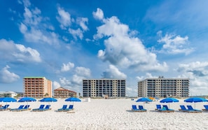 A beach with rows of blue sun loungers and umbrellas, a hotel with a green roof, and a large building with multiple balconies. - Island Wind West 177 Next to Hangout Pool Beach (Gulf Shores)