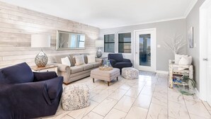 Un salon moderne avec un canapé beige, un fauteuil bleu foncé et un pouf de couleur claire. On y trouve une table basse en verre agrémentée d’objets décoratifs, une lampe sur une table d’appoint et un mur d’accent en bois.