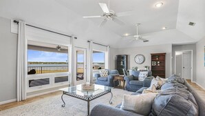 A spacious living room with a large gray sofa, a glass coffee table, and a view of the outdoors through large sliding doors. - Lagoon Time Beach Home- Elevator Beach Access (Gulf Shores)