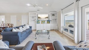 A spacious living room with a blue sofa set, a glass coffee table, and a view of the outdoors through large windows. - Lagoon Time Beach Home- Elevator Beach Access (Gulf Shores)