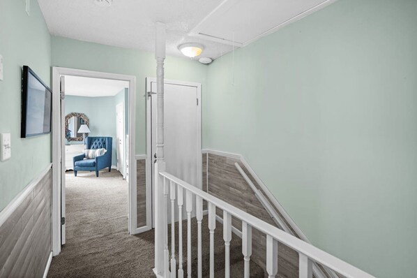 A hallway with a carpeted floor, a white railing, and a door leading to a room with a blue chair and a lamp. - Gulfside Townhome 32 Beachfront Townhouse w Pool (Gulf Shores)