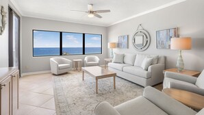 A spacious living room with a large window, a ceiling fan, a white sofa, a coffee table, and a side table with a lamp. - Four Seasons 502-e Pier Newly Remodeled (Orange Beach)