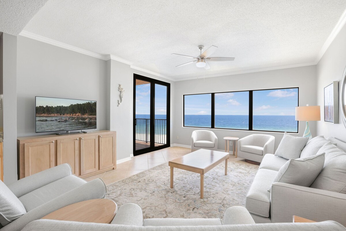 A modern living room with a large TV, comfortable seating, a ceiling fan, and a view of the ocean through large windows.