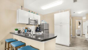 A modern kitchen with white cabinets, a granite countertop, and a built-in microwave. There are three blue bar stools and a coffee maker on the counter. - Grand Caribbean Pool Gym Beach View (Orange Beach)