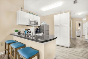 Una cocina moderna con armarios blancos, una encimera de granito y un microondas integrado. Hay tres taburetes azules y un refrigerador.