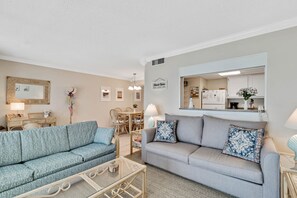 A living room with two sofas, a glass coffee table, a dining area with a table and chairs, and a kitchen visible through an open doorway. - Gulf Tower 12A Direct Gulf-front 2 2 Sleeps 6 (Gulf Shores)