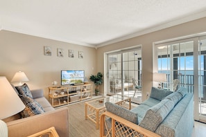 A spacious living room with a sofa, a television, and a large window with a view. - Gulf Tower 12A Direct Gulf-front 2 2 Sleeps 6 (Gulf Shores)