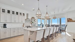 A modern kitchen with white cabinetry, a central island with bar stools, pendant lighting, and a view of the ocean through large windows. - Lil Tip Sea 8 BR Private Pool Elevator Beach (Gulf Shores)