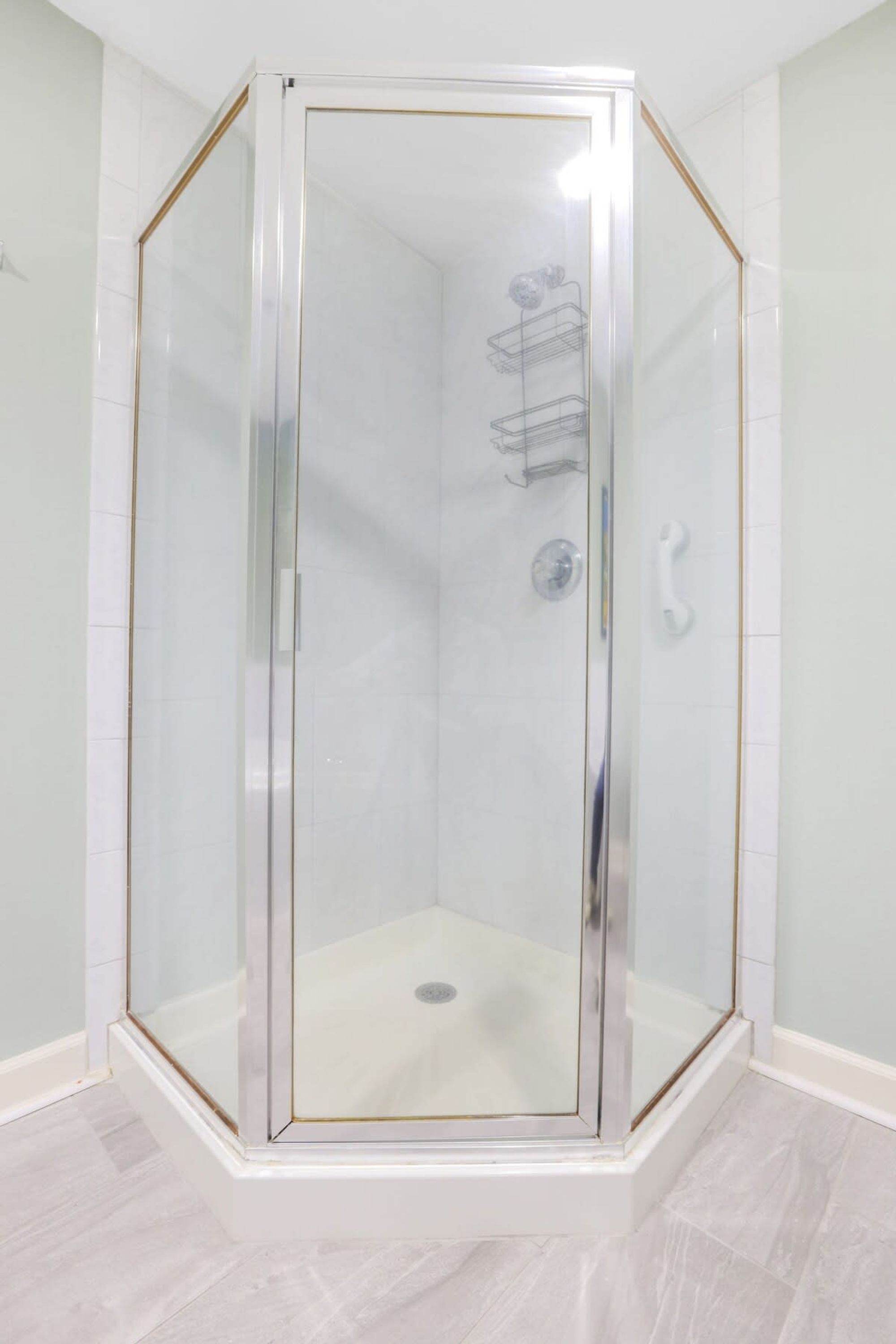 A glass-enclosed shower with a white tiled floor and walls, a metal shelf, and a white door handle.