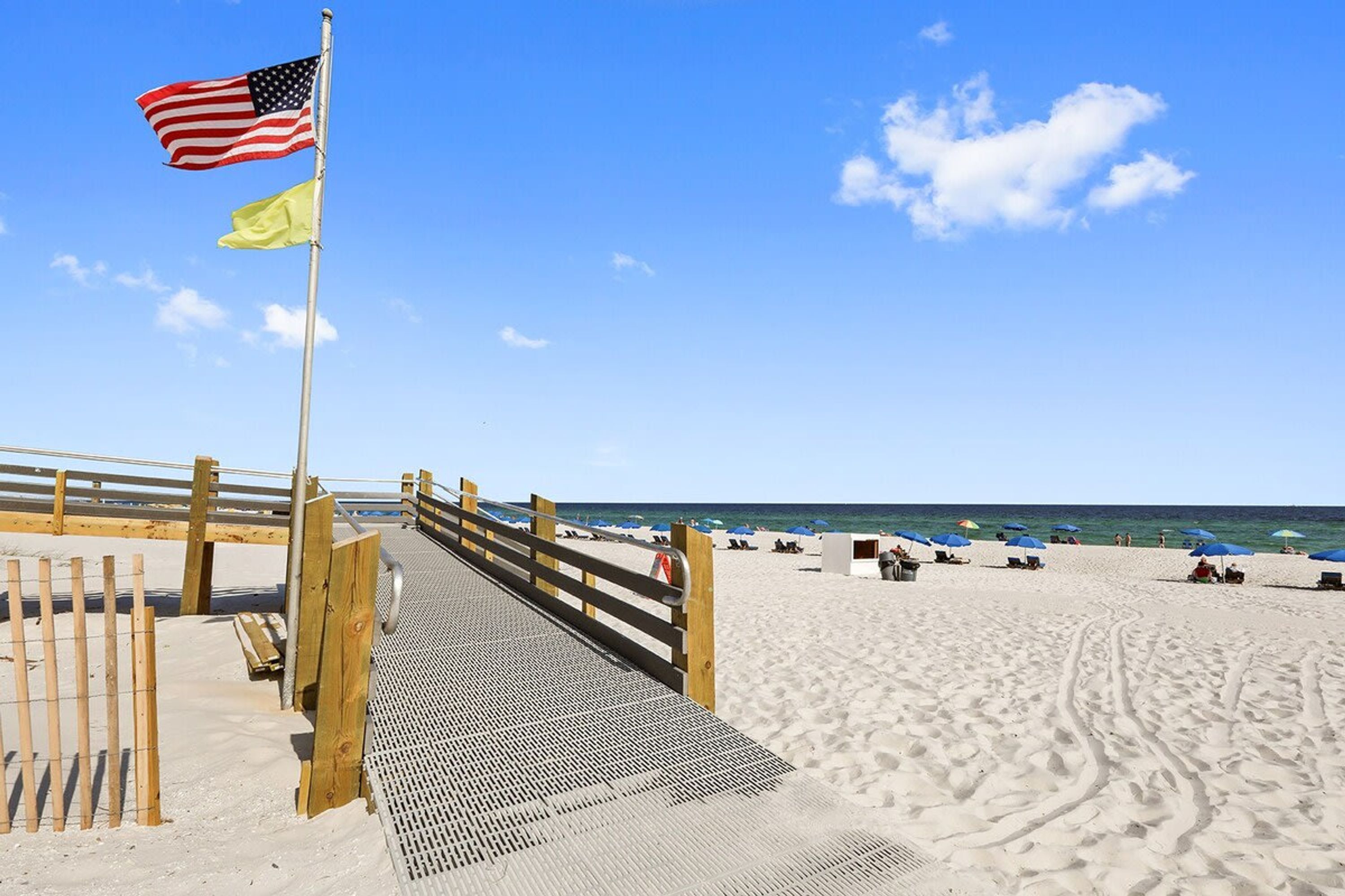 A wooden boardwalk leading to a beach with a flagpole displaying the American flag and a yellow flag.