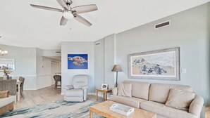 Een woonkamer met een beige bank, een salontafel, een plafondventilator, een ingelijst schilderij en een blauw vloerkleed.