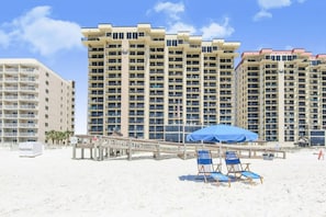 A beachfront hotel with multiple high-rise buildings, a sandy beach, and a wooden boardwalk with beach chairs and umbrellas.