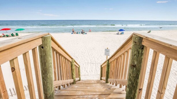 Eine Holztreppe führt zu einem Sandstrand mit Sonnenschirmen und Personen in der Ferne.