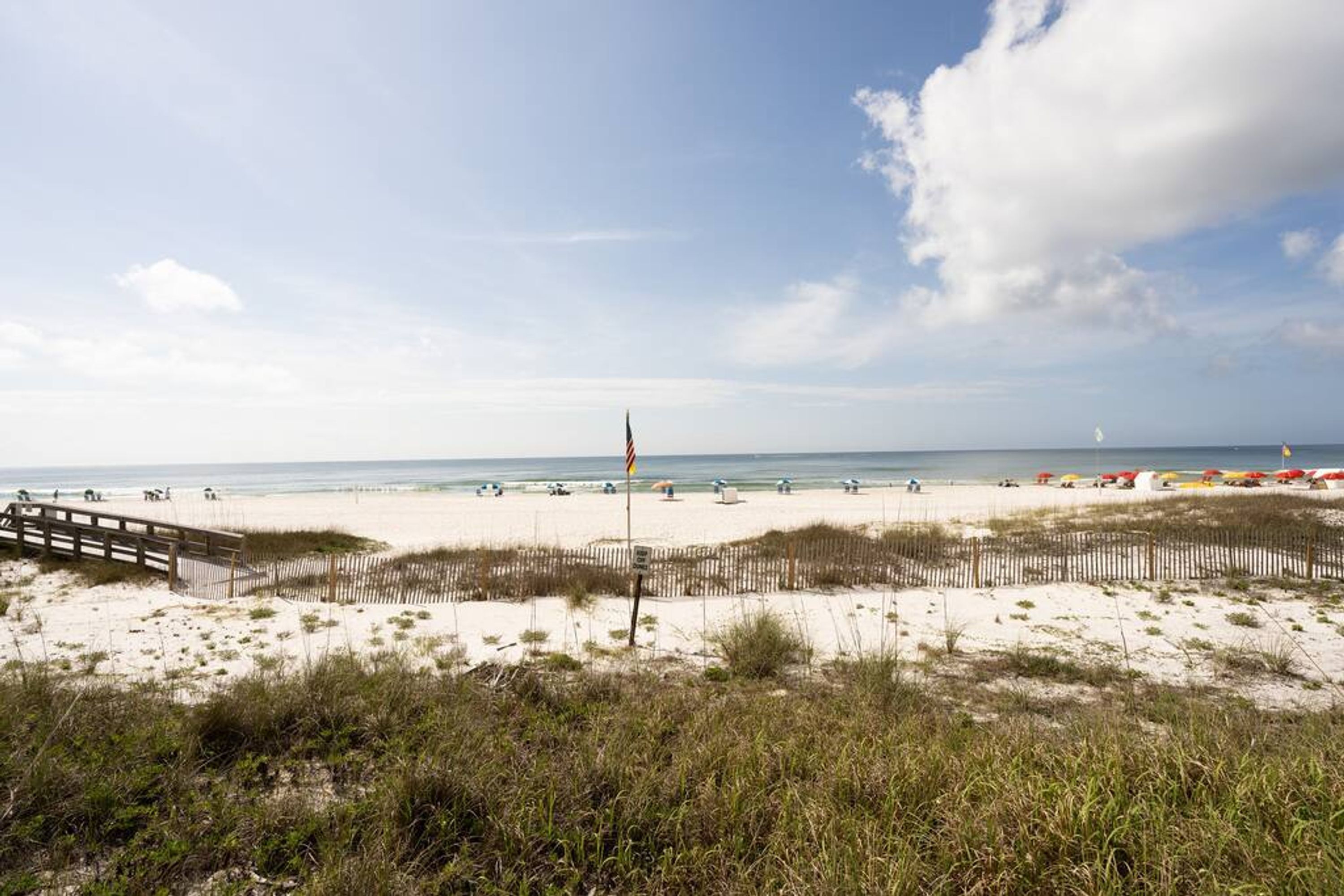 A beach with a wooden walkway, beach umbrellas, and a flagpole.