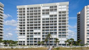 A multi-story hotel with a modern design, featuring balconies on each floor, located near a beach with a boardwalk and palm trees. - Pelican Pointe1204 3BR 2BA Gulf-front (Orange Beach)
