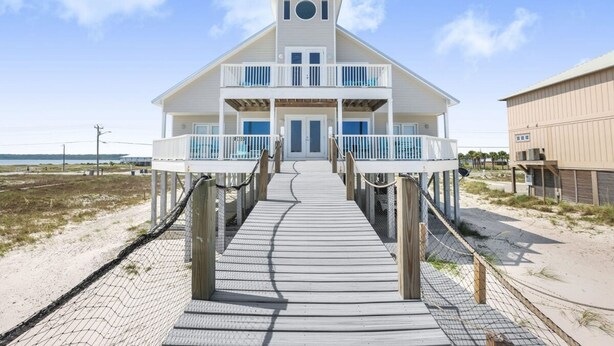 Une maison de plage à deux étages, dotée d’une terrasse en bois menant à la porte d’entrée, entourée d’une plage de sable et surplombant un ciel dégagé.