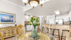 A dining area with a round glass table, chairs, and a vase with flowers. There is a kitchen in the background with white cabinets and a refrigerator. - Spanish Key 504 Next to Flora-bama Sleeps 9 (Pensacola)