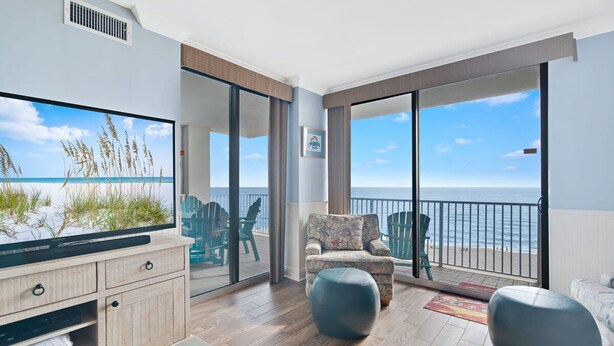 A modern living room with a large TV, wooden flooring, a sofa, a chair, and a balcony with a view of the ocean.