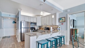 A modern kitchen with white cabinets, a granite island, teal bar stools, and pendant lighting.