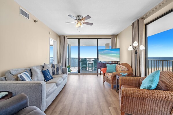 A spacious living room with a large TV, comfortable seating, a ceiling fan, and a balcony with a view of the ocean. - Shoalwater 906 3 3 Gulf-front Corner-unit (Orange Beach)