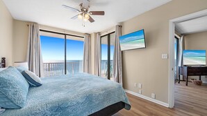 Un dormitorio con una cama, ventilador de techo, un gran ventanal con cortinas y un cuadro en la pared.