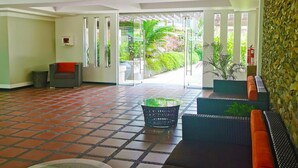 En rymlig entréhall med ett kaklat golv, en receptionsdisk, en sittgrupp med en soffa och stolar samt utsikt över en frodig trädgård genom glasdörrar.