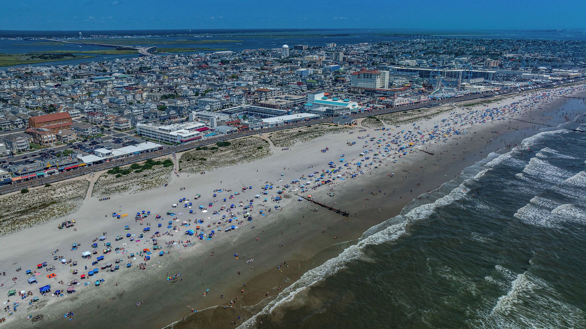 Vista aerea di una spiaggia con numerosi ombrelloni e persone, con una città sullo sfondo e un cielo sereno.