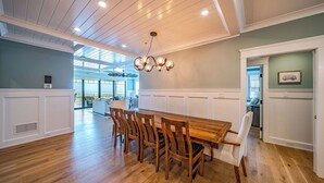 A spacious dining area with a wooden table and chairs, a modern chandelier, and a view of the outdoors through large windows.