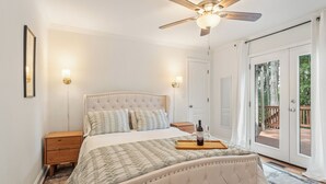 Een slaapkamer met een gestoffeerd hoofdeinde, een houten nachtkastje, twee wandlampen, een bed met een bedsprei met dessin, en een dienblad met een fles en glazen.