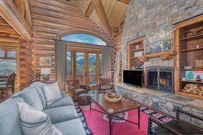 L’intérieur d’une cabane en rondins, avec une cheminée en pierre, des meubles en cuir et une grande fenêtre donnant sur les montagnes.