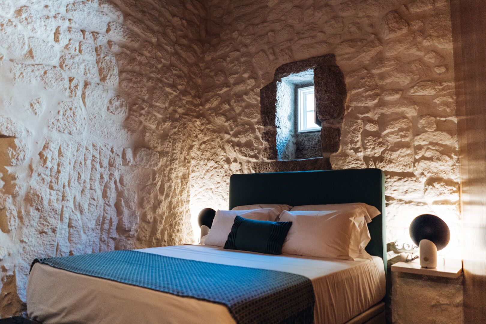 Una stanza con pareti in pietra, un letto, una piccola finestra e un comodino con una lampada.