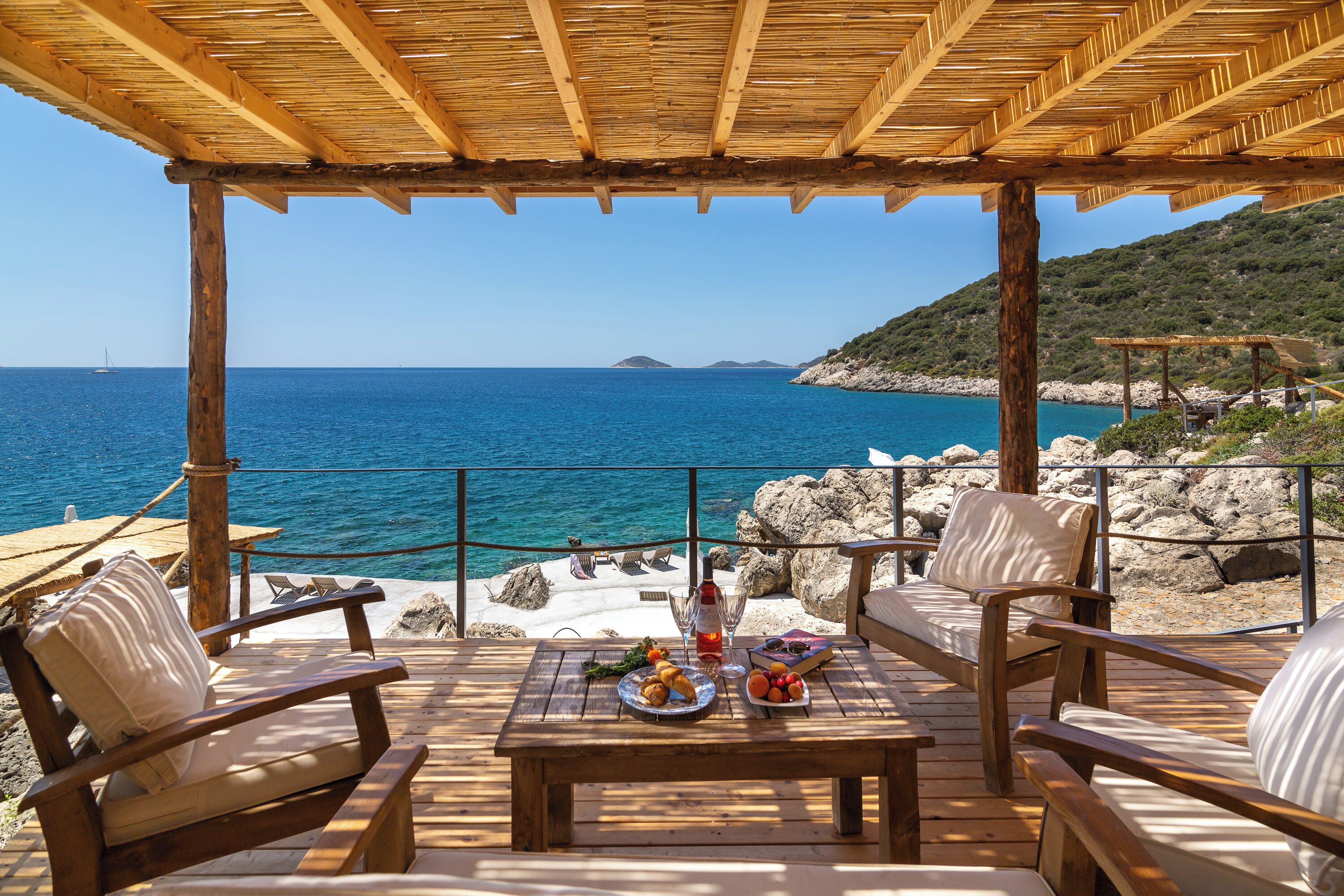 A wooden deck with a table set for two, overlooking a rocky coastline and the sea.