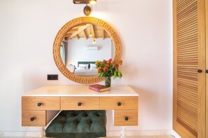 A wooden vanity with a mirror, a green upholstered stool, and a vase with flowers.