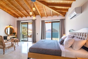 A bedroom with a wooden ceiling, a large bed, a bedside table, a chair, a mirror, and a view of the sea.