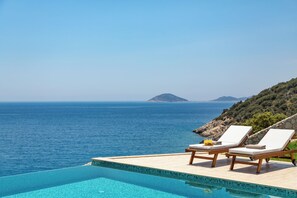A poolside area with two lounge chairs and a small table, overlooking a clear blue sea and a hilly landscape.