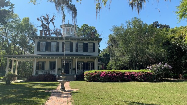 Historiallinen talo, jossa on keskeinen torni, tiilestä rakennettu polku, joka johtaa sisäänkäynnille, ja vehreää kasvillisuutta talon ympärillä.