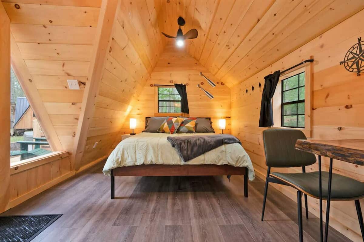 A cozy, wooden cabin bedroom with a bed, two bedside lamps, a chair, and a window.