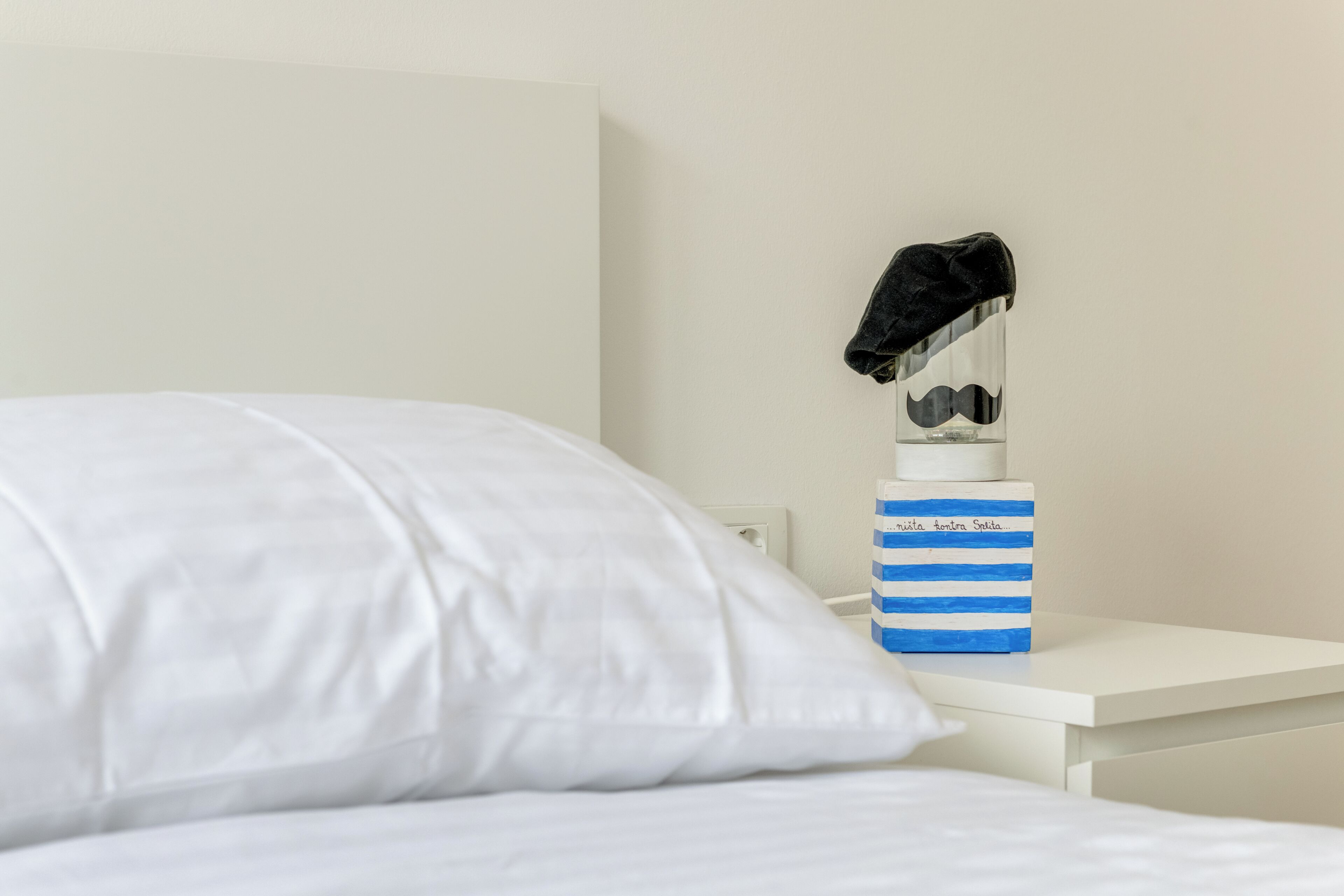 A neatly made bed with white sheets, a white nightstand with a blue-striped container holding a black hat and glasses, and a white wall.